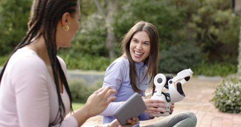 Young women laughing and sharing white companion robot while using tablet in park