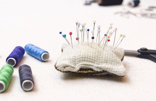 Colorful pins sticking on white cushion next to various colored threads and metal scissors on light surface. Perfect for illustrating sewing, needlework, crafts, DIY projects, textile art, and tailoring. Suitable for articles or websites about hobbies and handmade crafts.