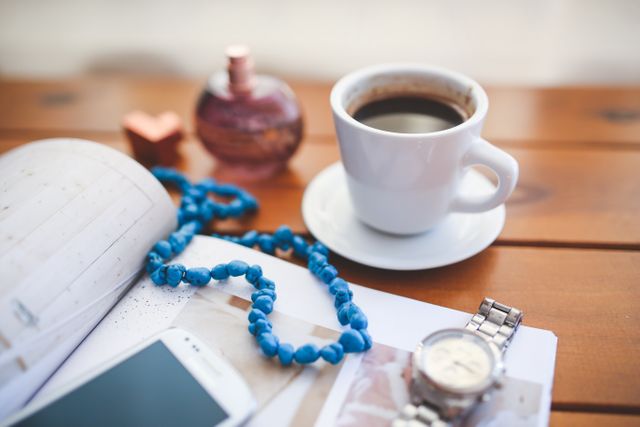 Cup of steaming coffee on wooden table surrounded by accessories including a watch, smartphone, bead necklace, and perfume bottle. Suitable for advertising relaxation, lifestyle, productivity, or morning routines.