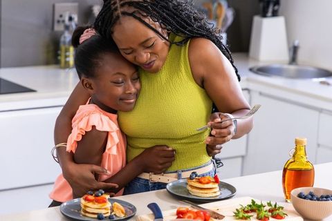 African american mother hugging daughter over homemade pancake breakfast in bright kitchen