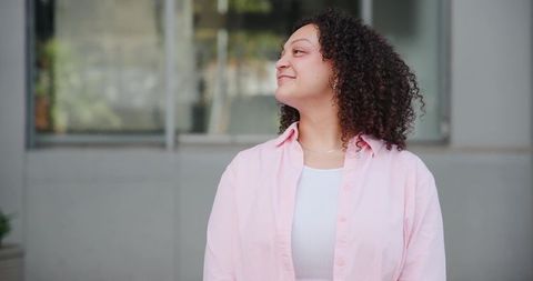 Serene young woman in pink shirt standing and looking up on urban facade with copy space