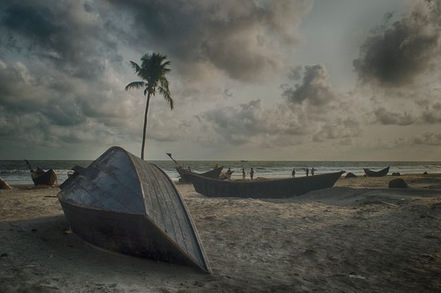 Fishing boats grounded on sandy beach with dramatic cloudy sky at dawn, offering serene view highlighted by a lone palm tree. Ideal for use in travel articles, nautical-themed projects, nature photography collections, and promotional materials for beach resorts.