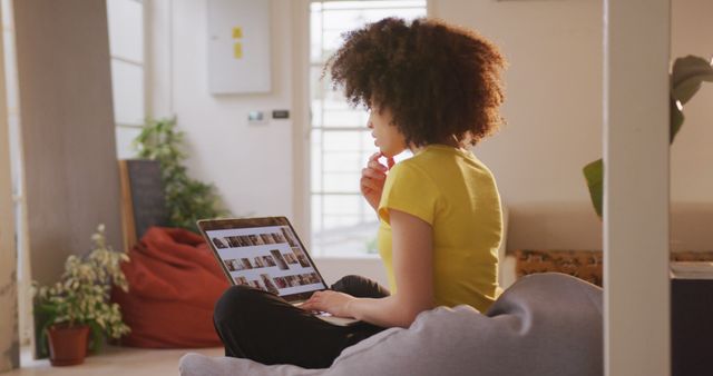 Young woman with an afro hairstyle is sitting comfortably in a cozy and modern home, browsing photos on a laptop. Plants and soft seating add to the relaxed atmosphere. Useful for articles on remote work, casual technology use, modern living at home, or lifestyle content featuring diverse individuals.