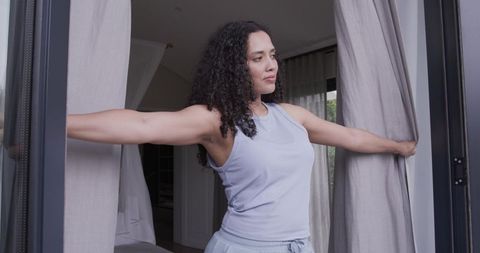 African american woman stretching and pulling curtains in bedroom doorway, morning light