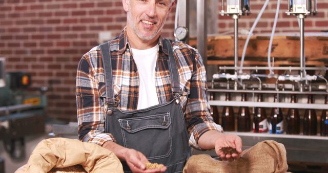 Male brewer in craft brewery holds malt and hops, smiling at camera. Background shows brewing equipment and brick wall. Ideal for use in articles, posts about craft brewing, beer production, artisanal food and drink industry, brewery business profiles.