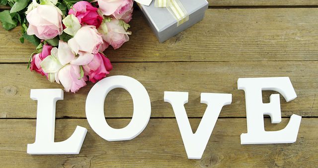 A romantic setting features the word LOVE in white letters, a bouquet of pink roses, and a gift box, with copy space. Perfect for expressing affection, this composition is ideal for Valentine's Day or an anniversary celebration.