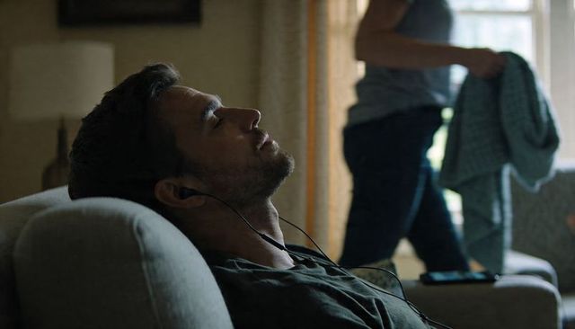 Man reclining on sofa listening to music with wired earbuds and smartphone on armrest