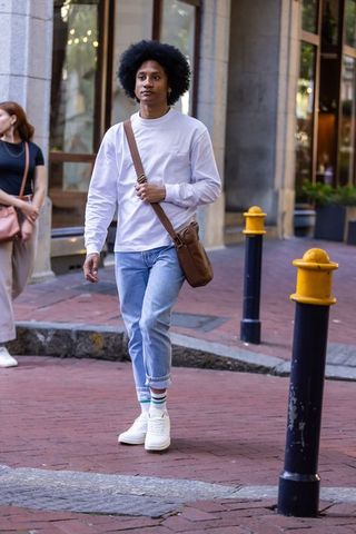 African american man walking on city sidewalk carrying brown crossbody bag in white shirt