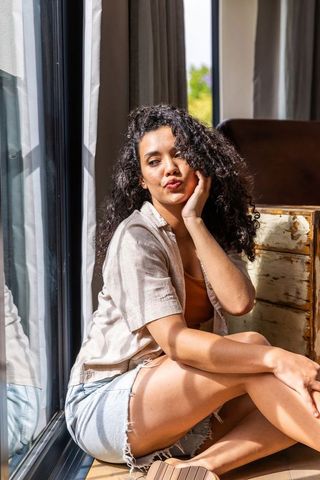 Sunlit african american woman sitting by glass door, resting cheek, wearing denim shorts
