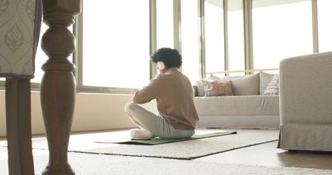 Man sitting cross-legged on green yoga mat in sunlit living room adjusting headphones