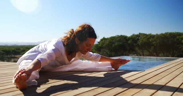 A young Caucasian woman practices yoga on a wooden deck overlooking a serene natural landscape, with copy space. Her focused expression and the tranquil setting suggest a moment of peaceful meditation and physical well-being.
