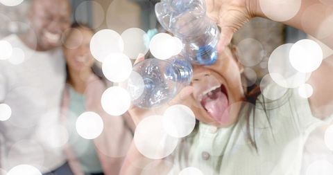 Playful child using water bottle binoculars laughing on couch with family bokeh lights