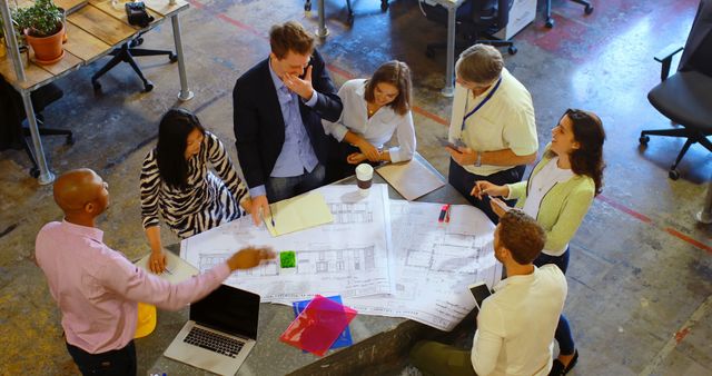 Diverse team collaborates over architectural plans in a modern office, actively discussing and brainstorming ideas. Useful for depicting teamwork, architecture, project planning, office environment, and corporate settings.