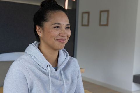 Smiling asian woman in grey hoodie sitting on bed with pearl earrings in minimalist bedroom