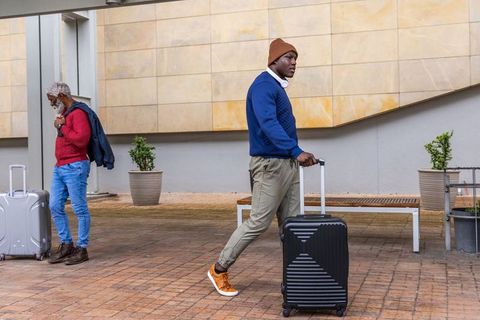 Young man pulling rolling suitcase under canopy walking with headphones and beanie