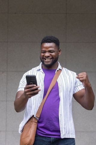 African man celebrating win checking phone wearing crossbody bag urban casual streetstyle