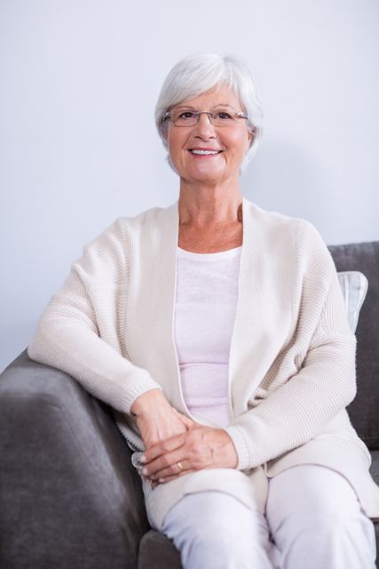 Senior woman with white hair and glasses sitting on a sofa in a clinic, smiling and looking relaxed. Ideal for use in healthcare, wellness, and medical contexts, such as brochures, websites, and advertisements promoting senior care, patient comfort, and medical services.