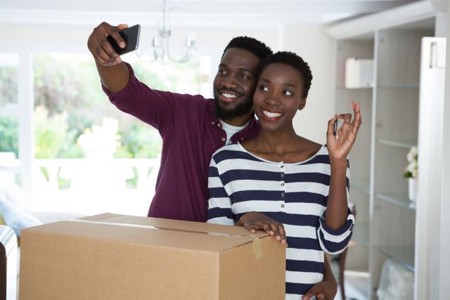 Happy couple taking a selfie while holding a new house key, standing next to a cardboard box. Ideal for use in real estate promotions, moving services advertisements, and homeownership articles.