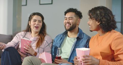 Diverse friends laughing and sharing popcorn on couch during cozy movie night at home