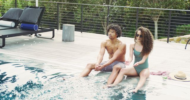 Image of happy diverse couple sitting at the side of swimming pool splashing water in the sun. Vacation, travel, happiness, relaxation and inclusivity concept.