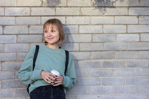 Young woman leaning on urban brick wall holding coffee cup wearing mint sweatshirt