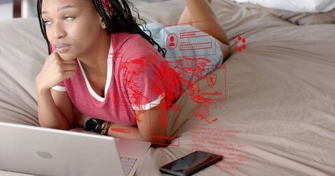 Young black woman lying on bed using laptop with holographic cybersecurity interface