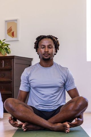 African american man meditating on green yoga mat at home holding remote, sunlit calm