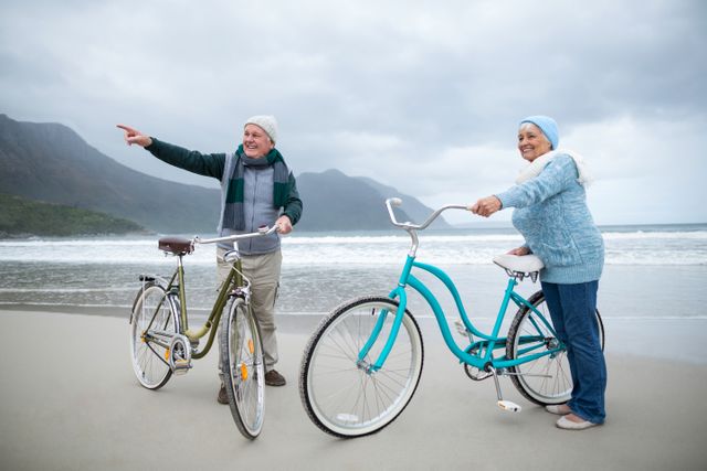 Senior couple enjoying a day at the beach with their bicycles, showcasing an active and healthy lifestyle. Ideal for use in advertisements promoting retirement plans, health and wellness products, travel and tourism, or outdoor activities. The scenic background with ocean and mountains adds a sense of adventure and tranquility.