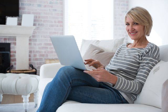 Mature woman sitting on a couch in a cozy living room, using a laptop and smiling. Ideal for themes related to home lifestyle, technology use, relaxation, and modern living. Suitable for articles, blogs, and advertisements focusing on mature audiences, home comfort, and digital connectivity.
