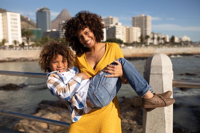This image captures a joyful moment between a mother and her son by the seaside promenade on a sunny day. The mother, wearing a yellow dress, holds her son in her arms as they both smile at the camera. The background features the ocean and a cityscape with buildings. This image is perfect for use in family-oriented content, travel promotions, lifestyle blogs, and advertisements focusing on family bonding and outdoor activities.