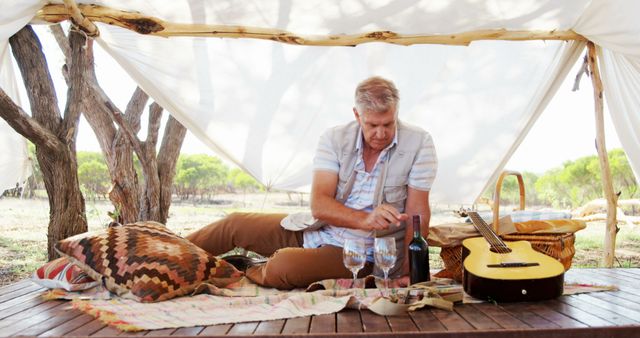 Mature man enjoying a serene moment outside under a canopy with a bottle of wine and an acoustic guitar. Perfect for promoting relaxation, outdoor activities, and leisure time. Ideal for use in travel brochures, lifestyle blogs, and outdoor activity promotions.