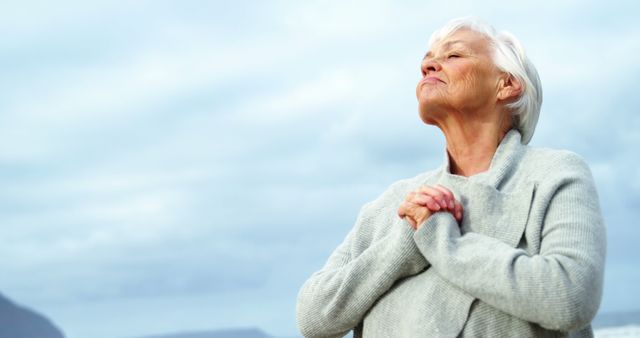 A senior Caucasian woman appears serene as she closes her eyes and clasps her hands in a peaceful outdoor setting, with copy space. Her expression suggests a moment of gratitude or reflection, adding a sense of tranquility to the image.