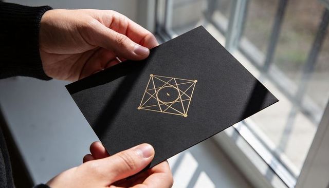 Hands holding luxury black invitation with gold geometric emblem by sunlit window