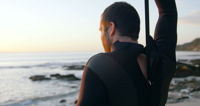 Back view of man zipping up wetsuit near ocean shore at sunset, ideal for content related to outdoor activities, water sports, surfing, beach lifestyle, and adventure. This photo can be used in fitness blogs, travel websites, advertisements for surf gear, or motivational content about embracing nature.
