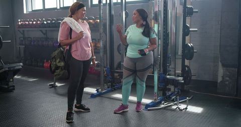 Two diverse women chatting after workout by squat rack in sunlit urban gym