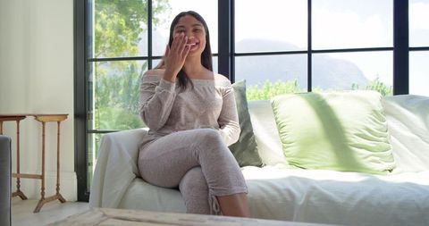 Smiling african woman relaxing on sunlit sofa in off-shoulder loungewear cozy home interior