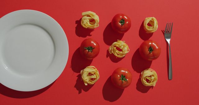 Image shows an empty white plate with fresh tomatoes and pasta nests on a vibrant red background. A fork sits nearby, adding to the food arrangement. Ideal for use in culinary blogs, cooking websites, food magazines, and restaurant advertisements highlighting Italian food or recipes.