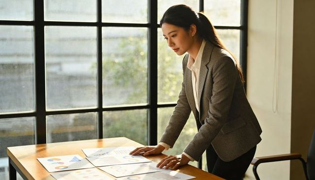 Asian businesswoman leaning over table reviewing financial charts by mullioned window