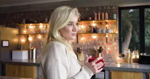 Blonde Woman Holding Red Mug in Cozy Brick Kitchen with Warm Ambient Lights
