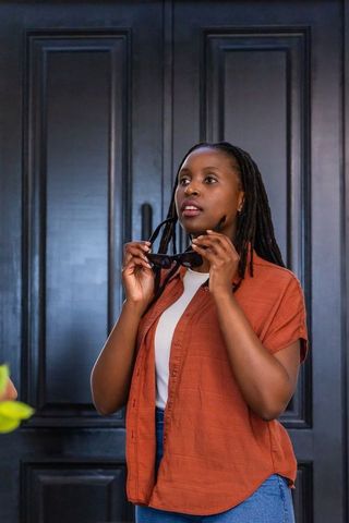 Pensive woman standing by dark paneled door wearing rust shirt holding sunglasses
