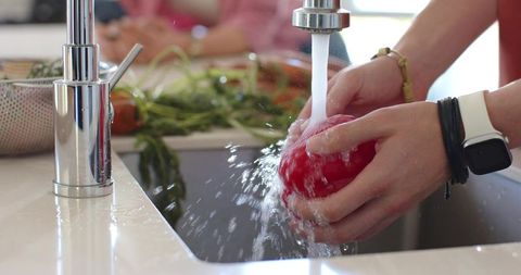 Closeup hands rinsing tomato under chrome kitchen faucet with smartwatch, greens and colander