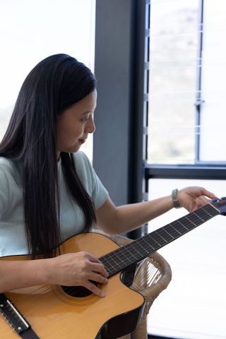 Mature african woman tuning acoustic guitar by window practicing relaxed home music