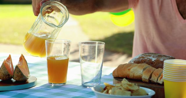 This image depicts a person pouring fresh orange juice into a glass during a sunny outdoor picnic. The table is covered with a green gingham tablecloth and is set with various food items including sliced bread, melon slices, and a bowl of chips. Great for depicting summer leisure activities, food and drink enjoyment, or promotional materials for picnic supplies and outdoor events.