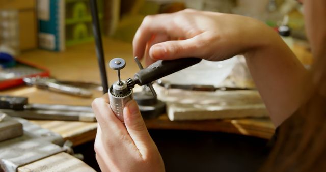 Close-up of a person's hands working with a rotary tool on a small object, a piece of jewelry, with a workshop environment in the background. Precision and craftsmanship are evident as the individual focuses on the intricate task at hand.