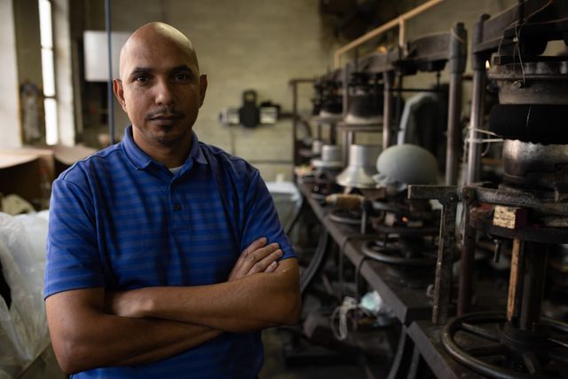 Portrait close up of a bald biracial man with crossed arms looking straight to camera in the workshop at a hat factory.