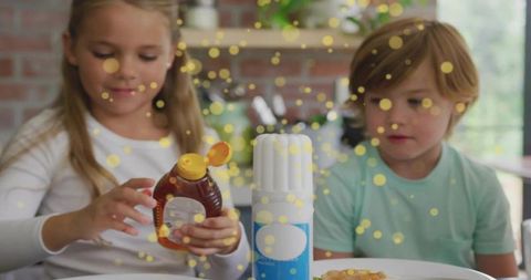 Girl pouring honey onto pancakes while brother watching in bright home kitchen morning