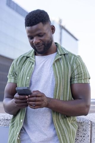 African american man checking smartphone while leaning on urban ledge wearing green shirt