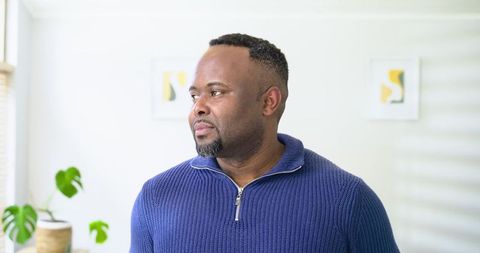 African American man standing by blinds looking out in bright interior wearing navy zip sweater