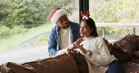 Diverse couple sharing cozy holiday moment on sofa wearing santa hat and reindeer headband