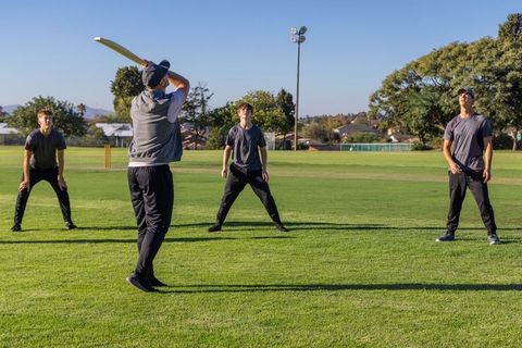 Male teammates playing cricket practice on sunny suburban pitch, batting, fielding, teamwork